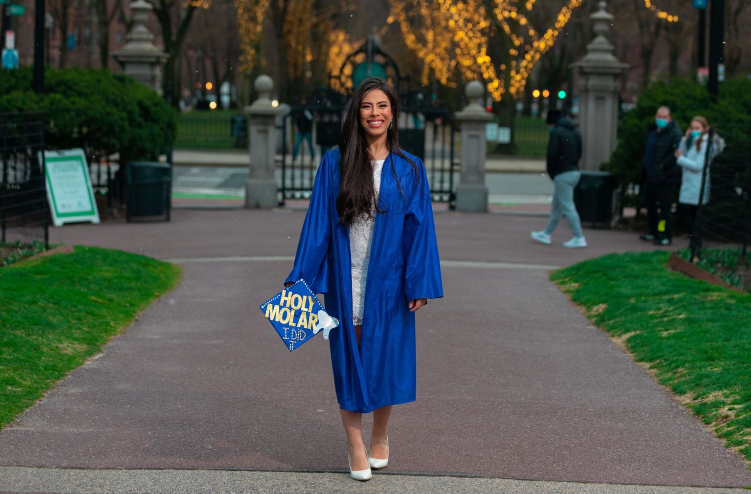 Dental school graduate walking on campus path in blue regalia holding decorated cap.