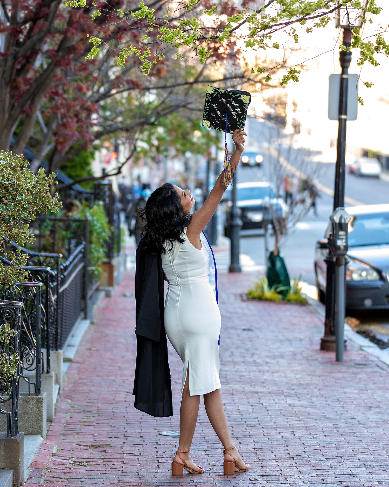 Graduate in white dress and cap raises arm triumphantly on historic brick sidewalk.