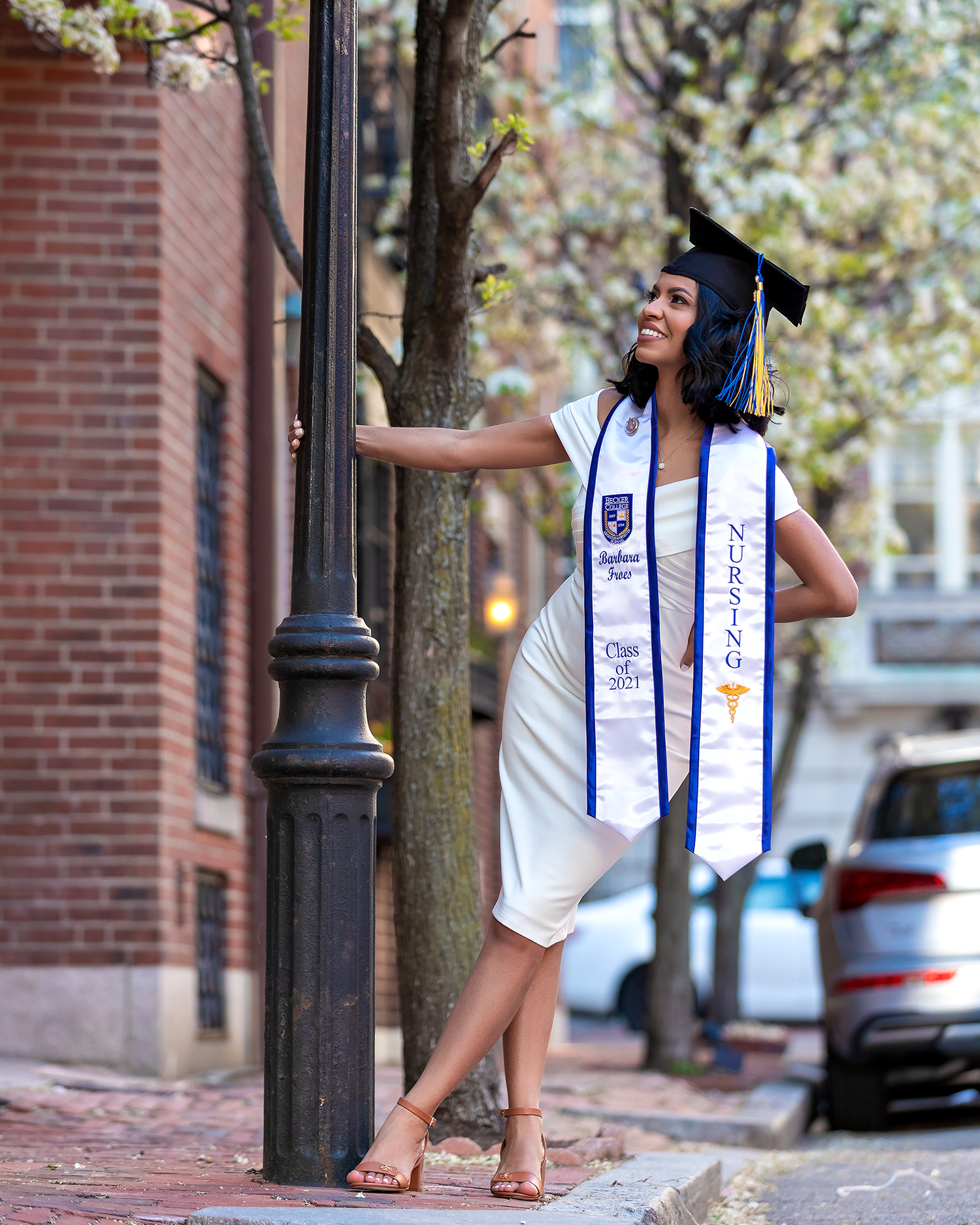 Graduate in white dress and blue sash poses joyfully against brick wall on graduation day.