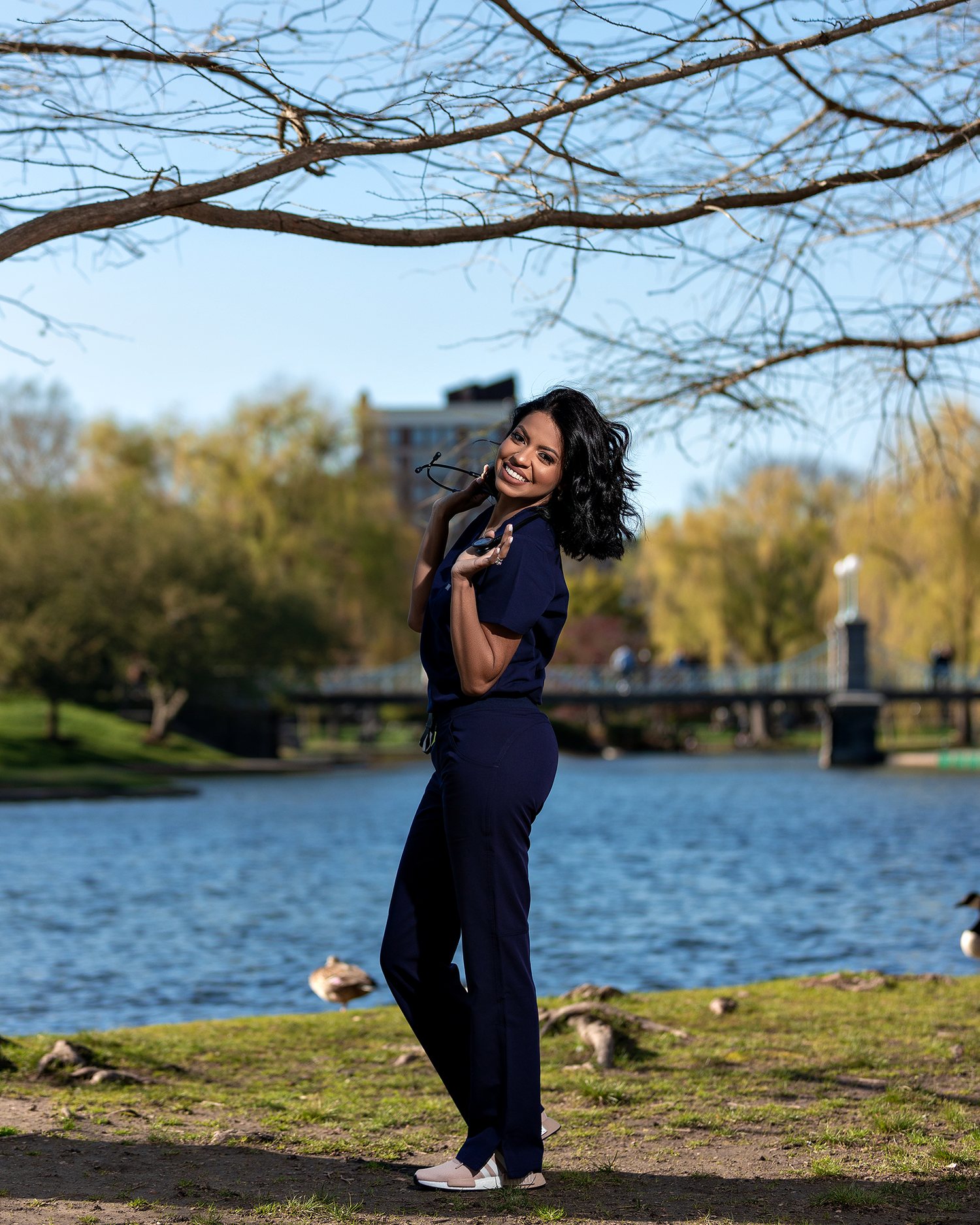 Graduate in navy outfit poses playfully by riverside with trees and bridge in background.