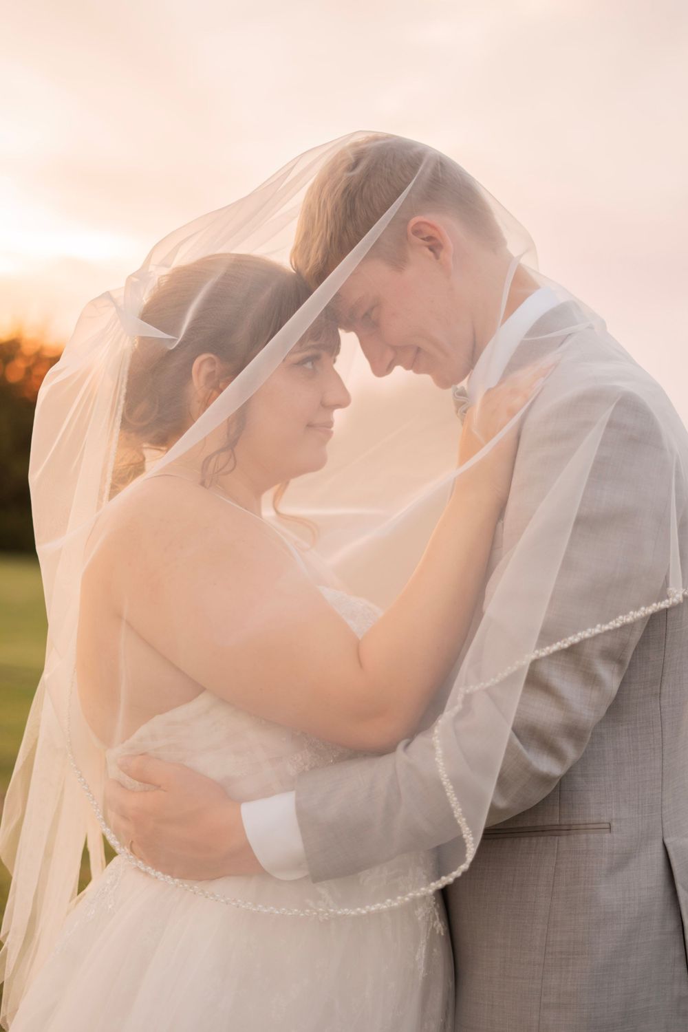 Newlyweds embrace tenderly under a bridal veil at sunset, foreheads touching in a romantic outdoor portrait.