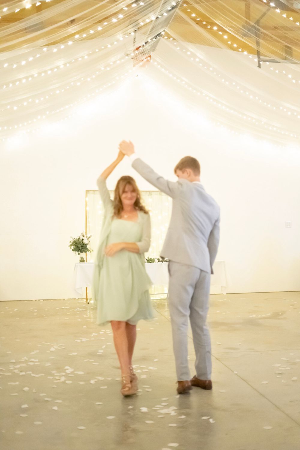 Groom twirls a woman in a mint green dress on a decorated dance floor under warm string lights.
