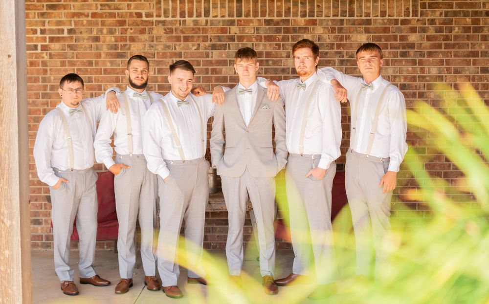 Groom and five groomsmen in gray suits with suspenders posing against brick wall at wedding.