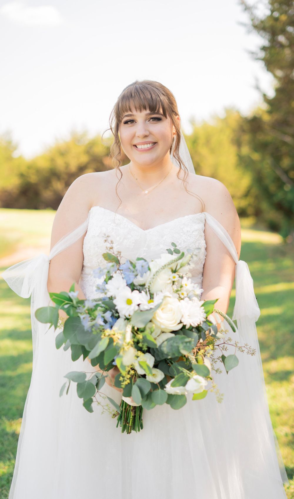 Smiling bride in strapless lace wedding gown holding a lush white and green bouquet outdoors.