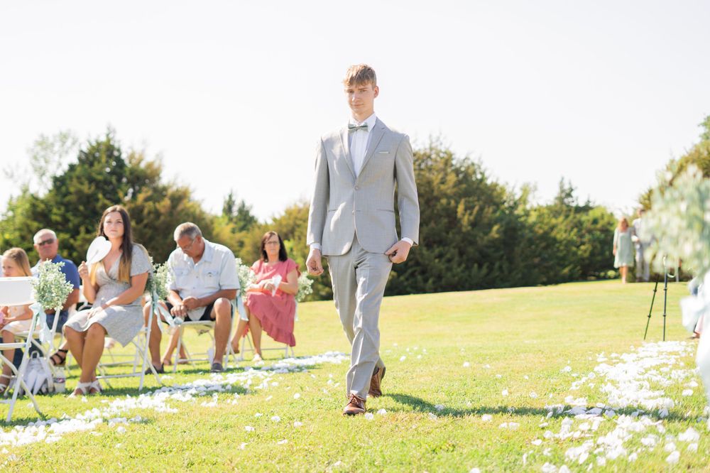 Groomsman in grey suit walking down the flower petal-lined aisle at a sunny outdoor wedding ceremony.