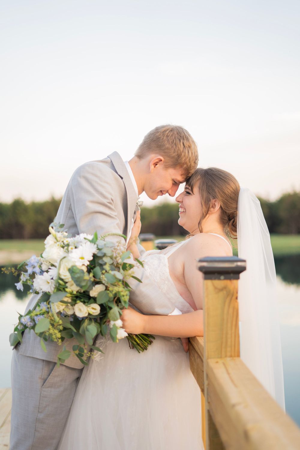 Smiling bride holds a white floral bouquet as she and groom touch foreheads on a lakeside wooden dock.