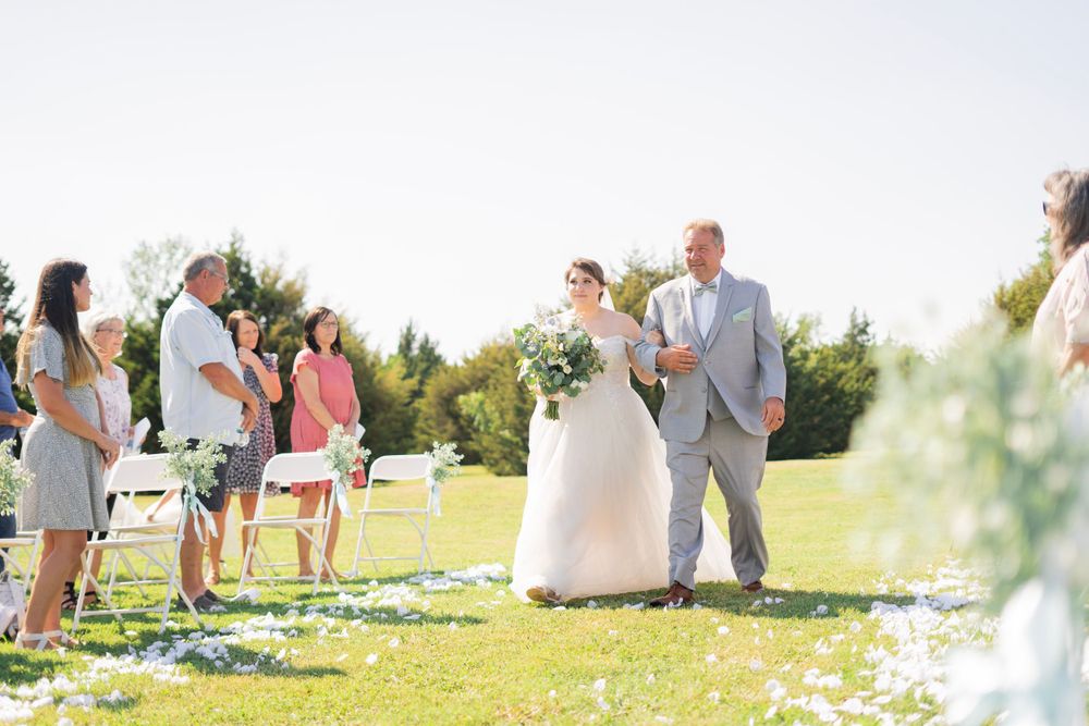 Father walking bride down the flower petal-lined aisle at a sunny outdoor wedding ceremony.