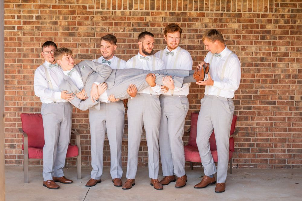 Groomsmen playfully carry the groom in gray suits against a rustic brick wall backdrop.
