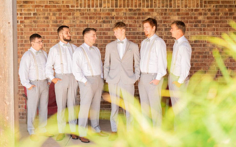 Groom and groomsmen in gray suits and suspenders chatting casually against rustic brick wall outdoors.