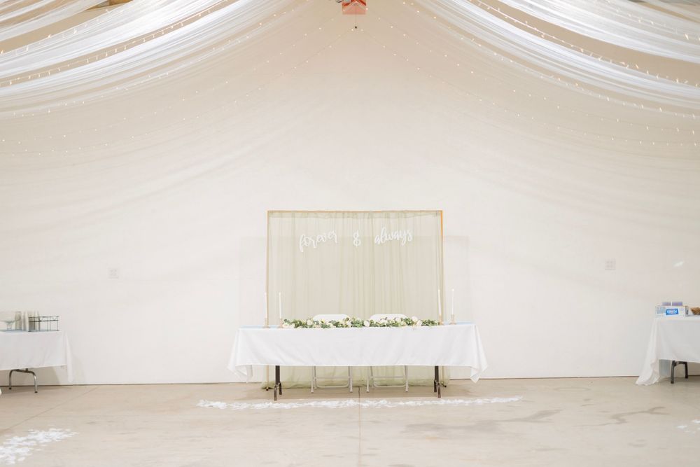 Elegant wedding sweetheart table with white draping backdrop and greenery garland in a white tent venue.