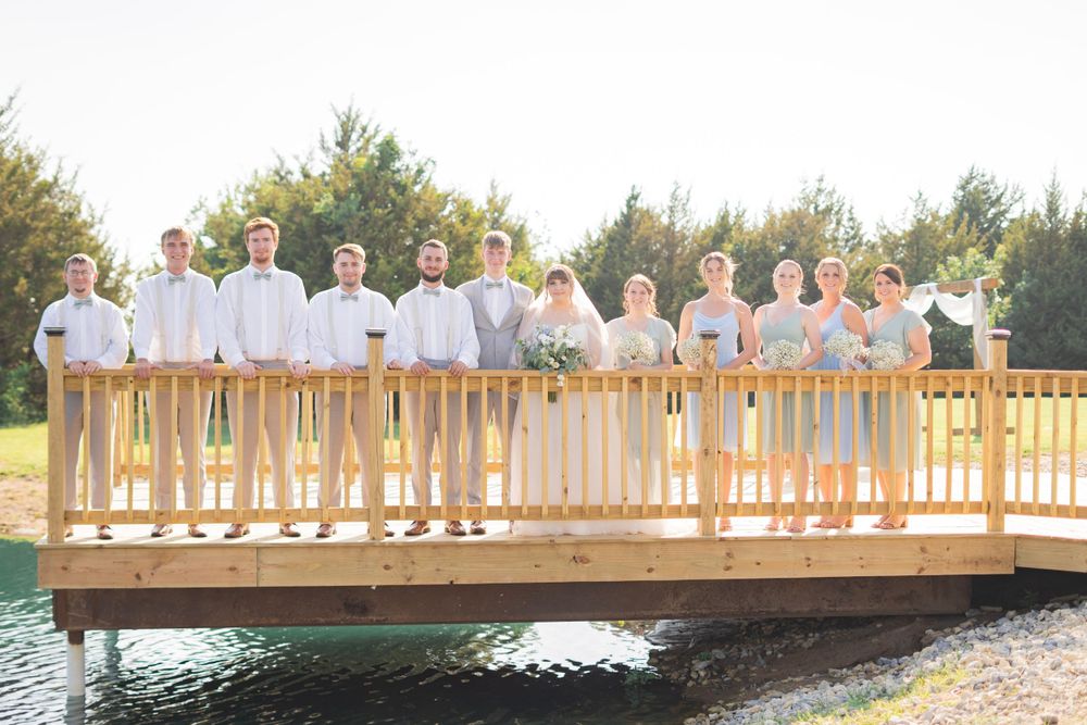 Wedding party posing formally on a wooden bridge, groomsmen in grey suits and bridesmaids in blue dresses.
