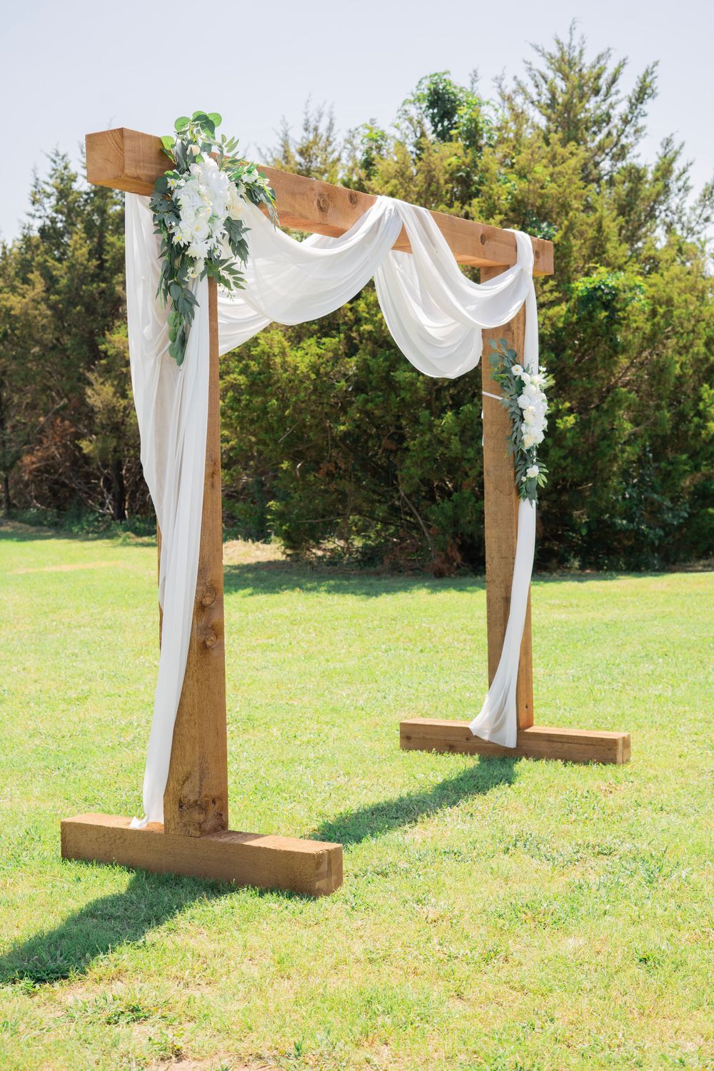 Rustic wooden wedding arch decorated with white fabric draping and white floral greenery on green lawn.