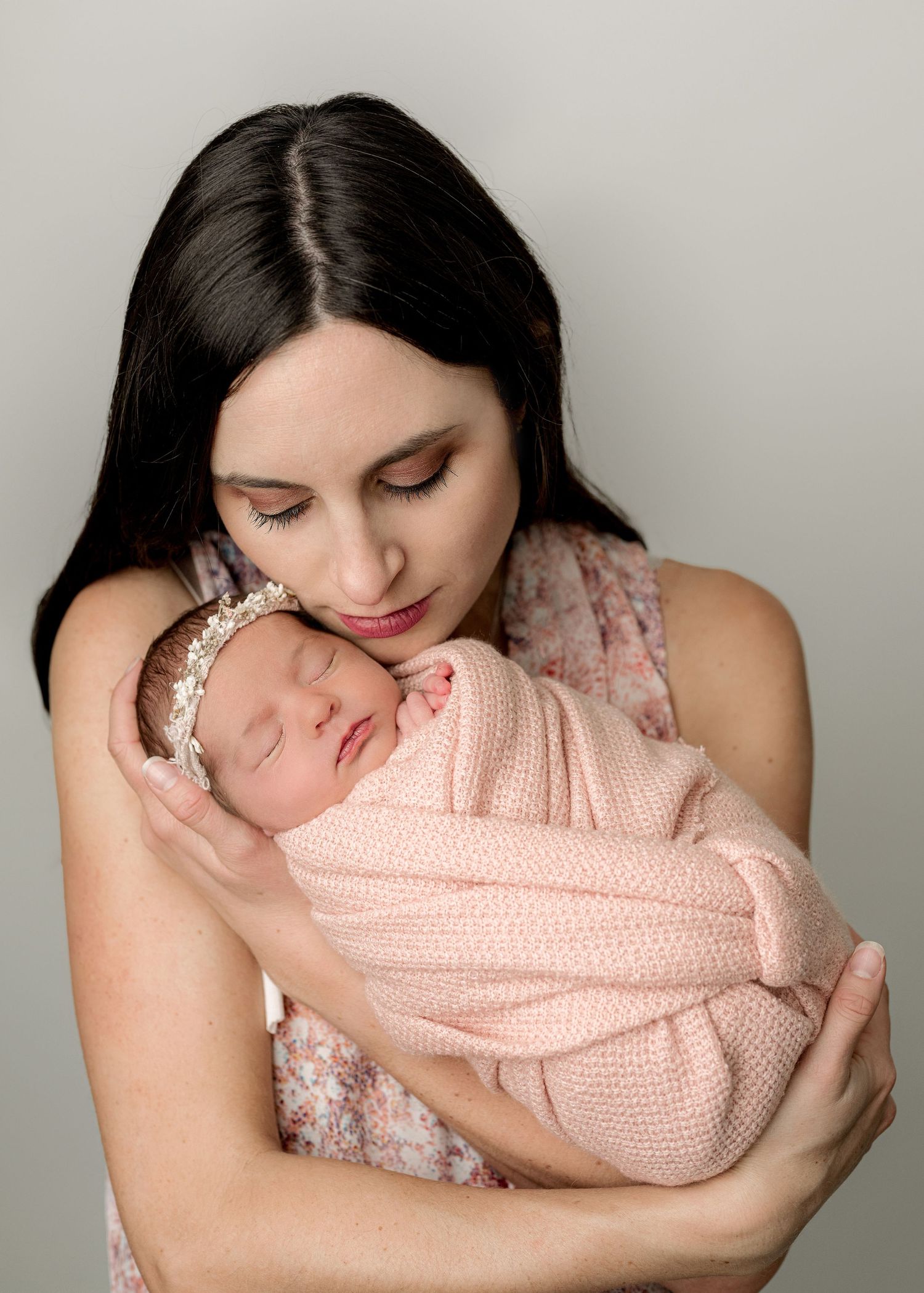 A tender moment between mother and newborn baby wrapped in pink knit fabric against a gray background.