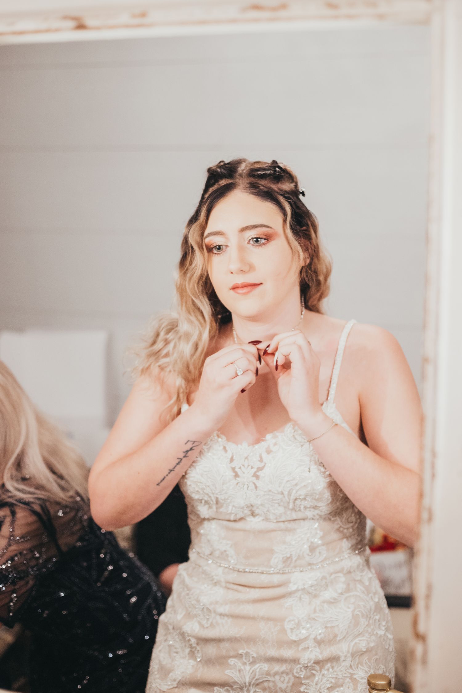 Someone wearing a white beaded wedding dress adjusts their necklace while getting ready in a softly lit room.