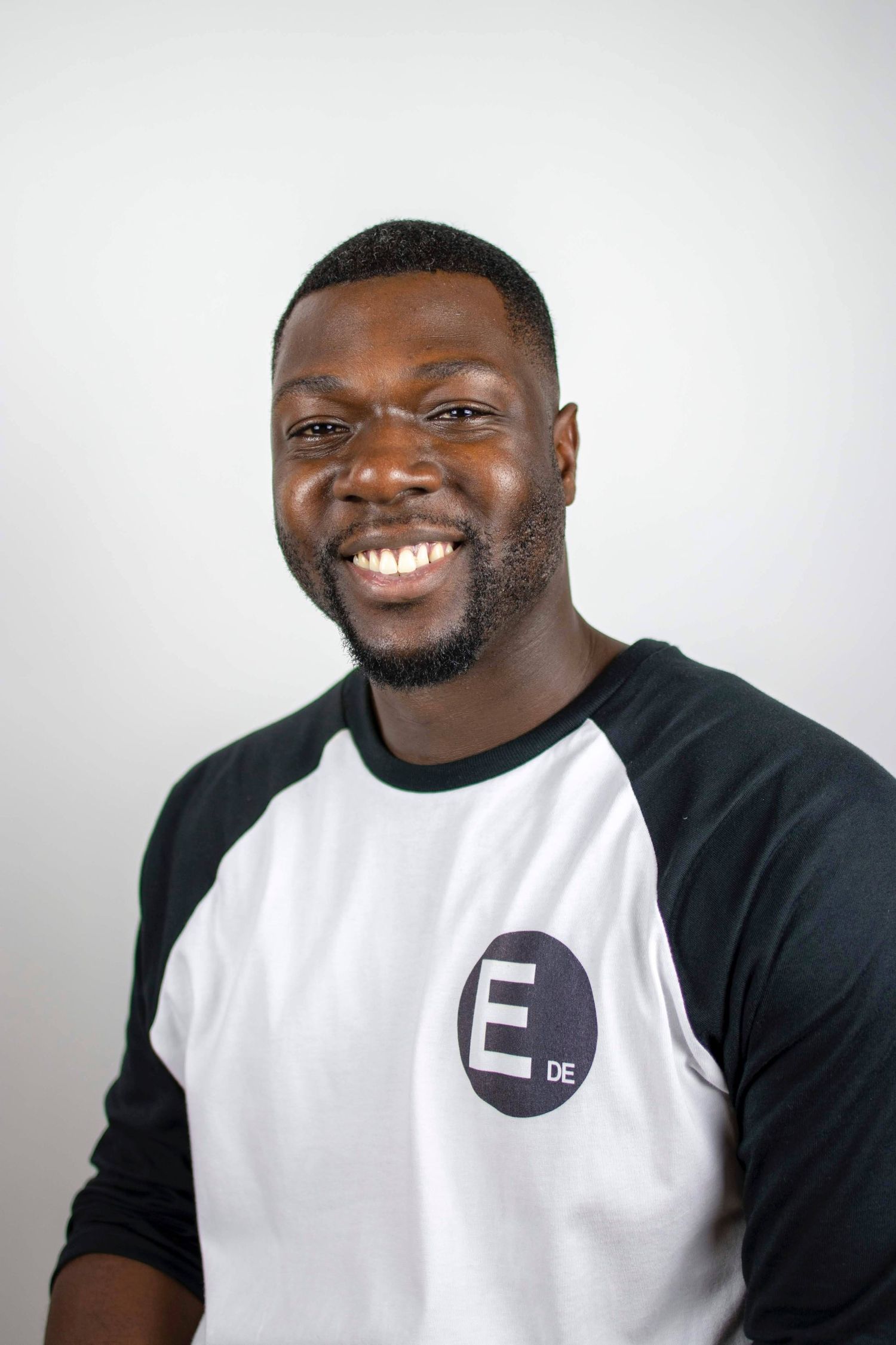 A person wearing a black and white baseball-style shirt with an 'E' logo smiles warmly against a light background.