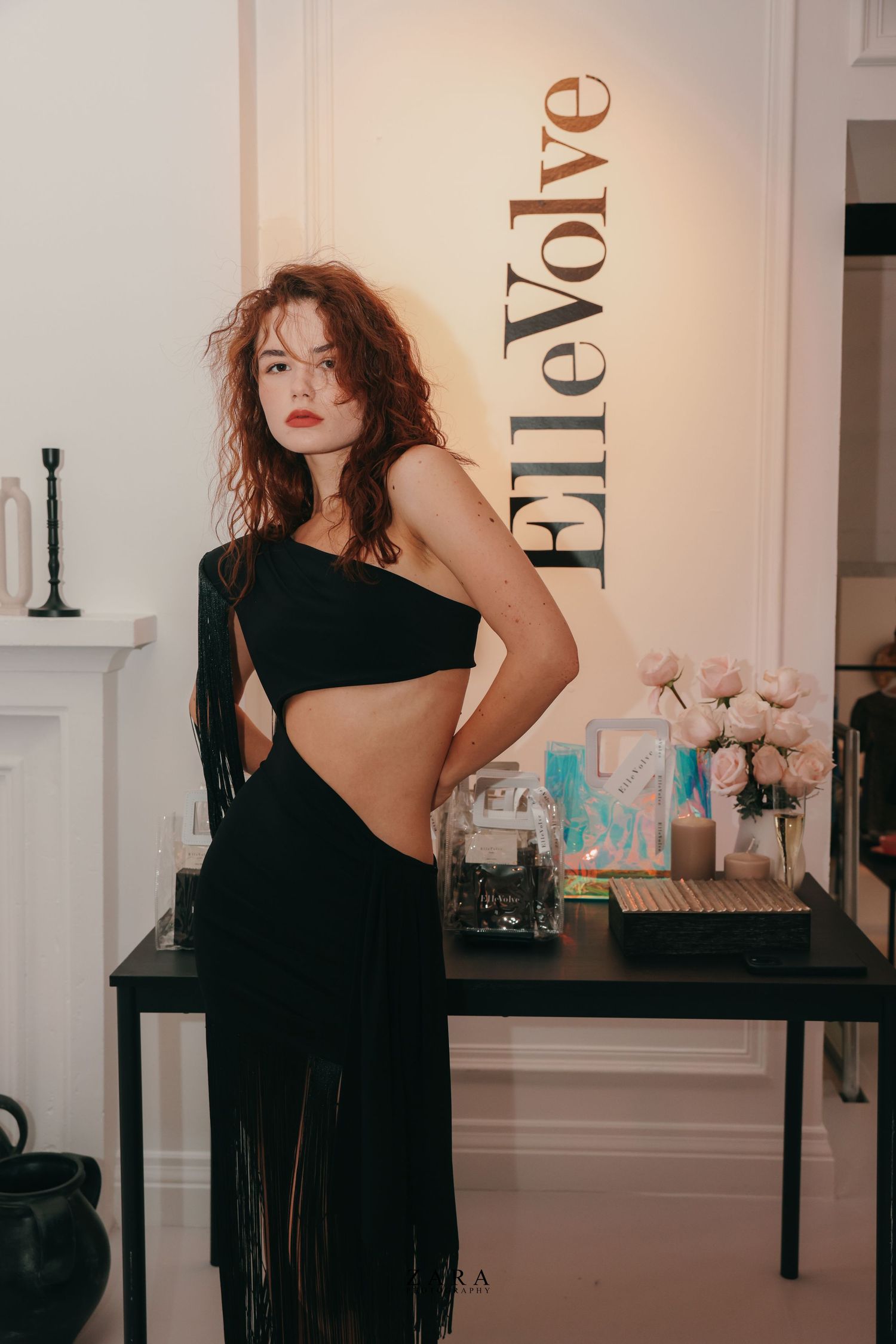 Fashion model in black outfit posing against wall with text branding.