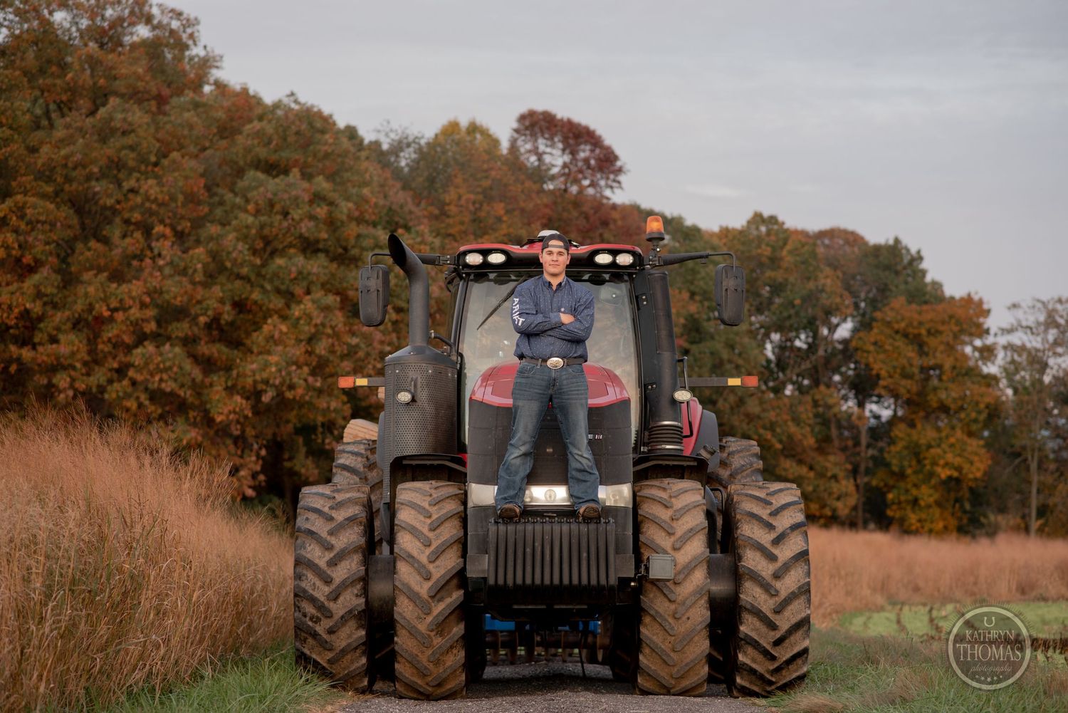 Silhouette of person standing atop large red tractor against autumn forest backdrop.