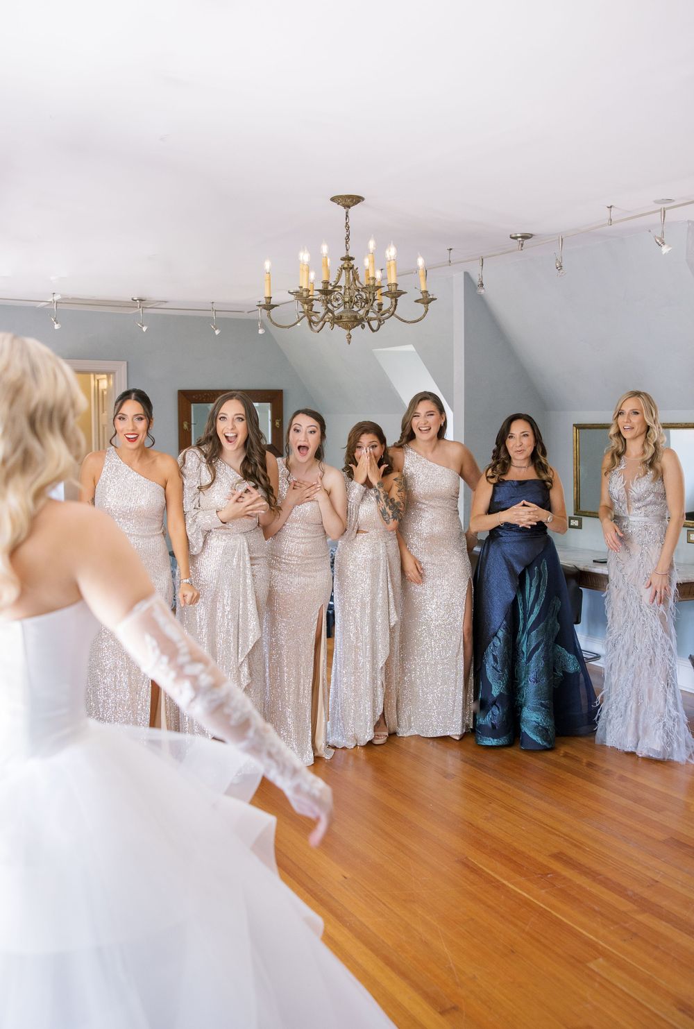Bridesmaids in sparkly gowns gathered in room with chandelier watching bride in white dress.