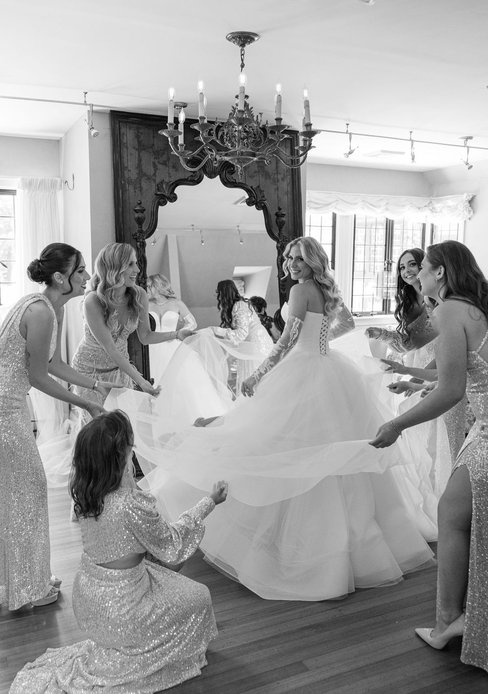 Bridesmaids helping with a flowing white wedding dress in an elegant room with chandelier.