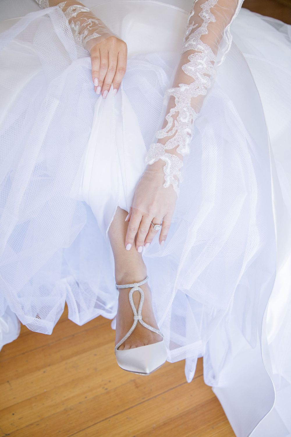 Detail shot of bride's lace sleeves, white shoes and flowing dress hem on wooden floor.