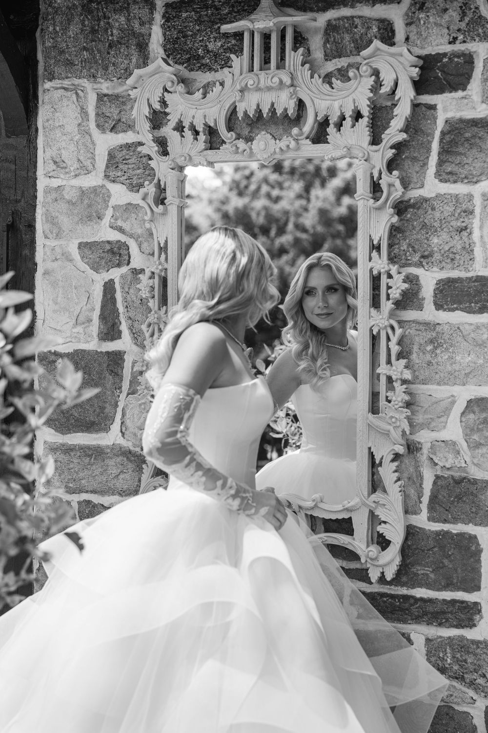 Black and white photo of wedding dress reflection in ornate mirror against stone wall.