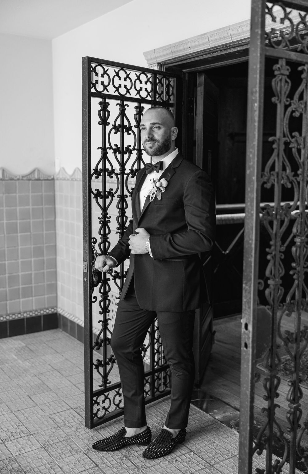 Black and white portrait of groom in tuxedo standing by ornate iron gate.