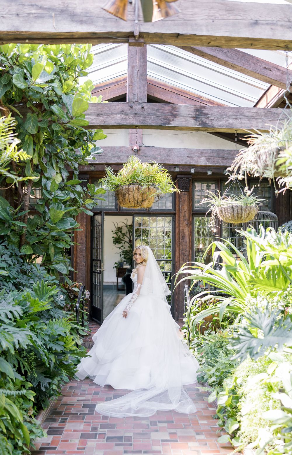 Bride in flowing white gown poses in lush garden setting with wooden beams overhead.