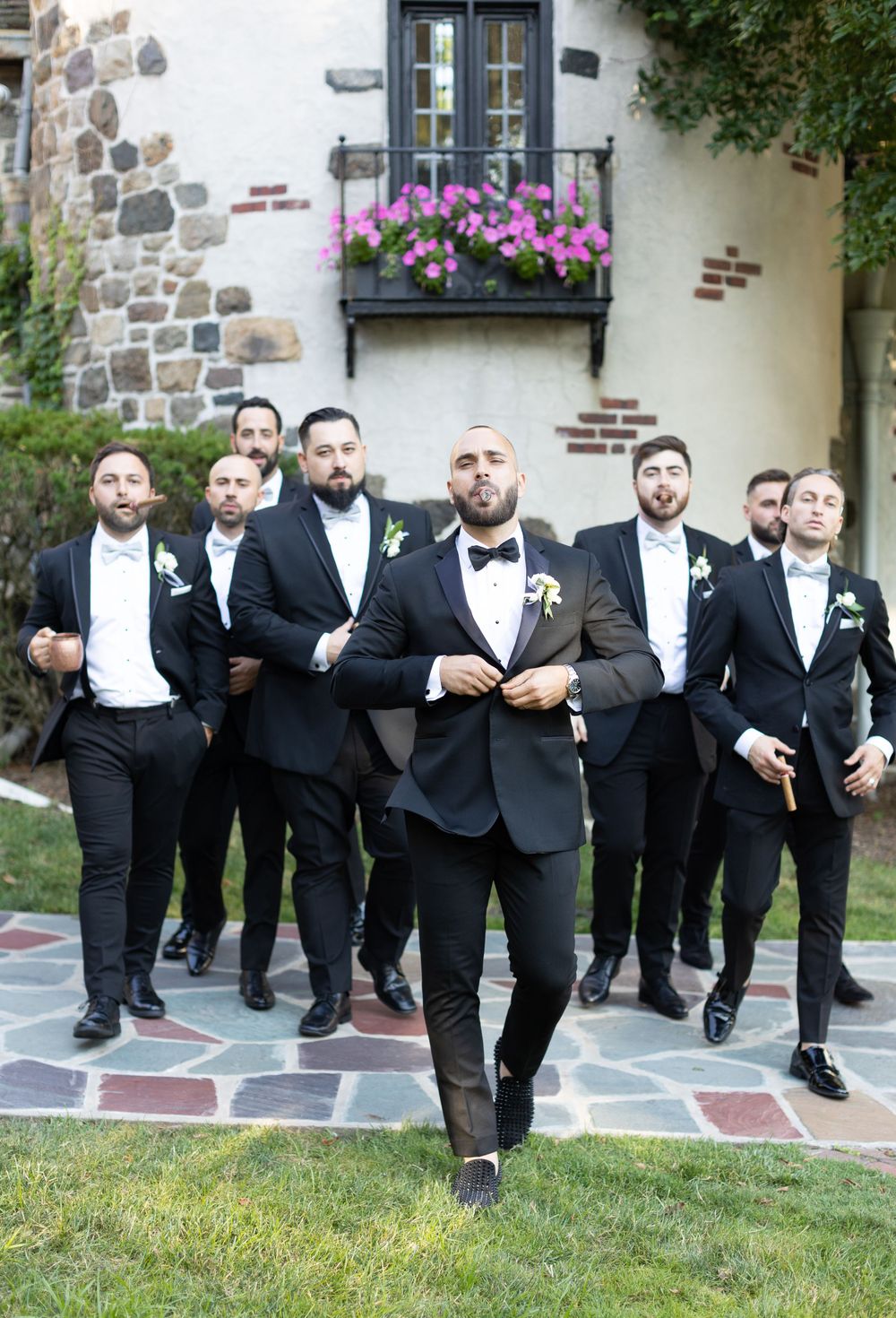 Groomsmen in black tuxedos pose in front of stone building with flower boxes.