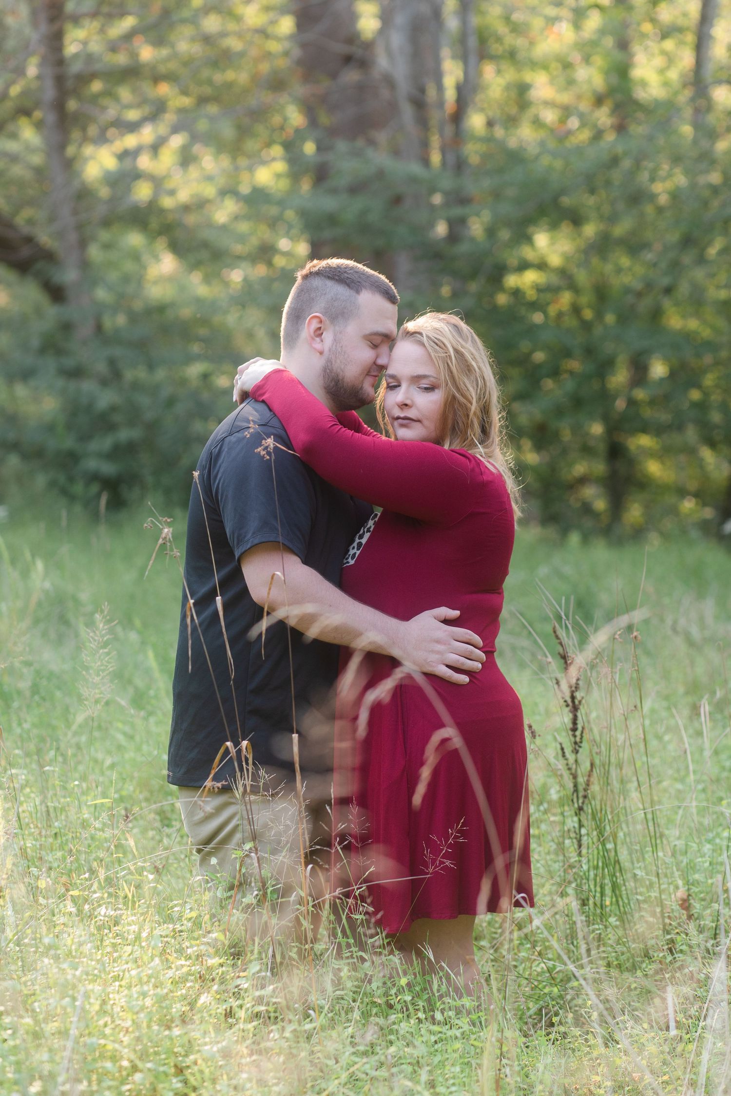 A romantic photoshoot in a sunlit meadow with tall grass and trees in the background.