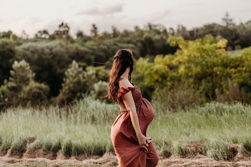 A woman in a rust-colored dress stands in a sunlit meadow during golden hour.