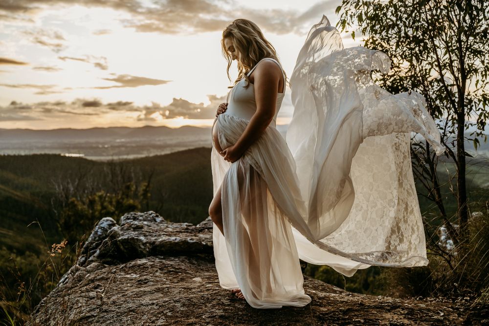 A flowing white dress billows in the wind during a mountaintop maternity photo shoot at sunset.