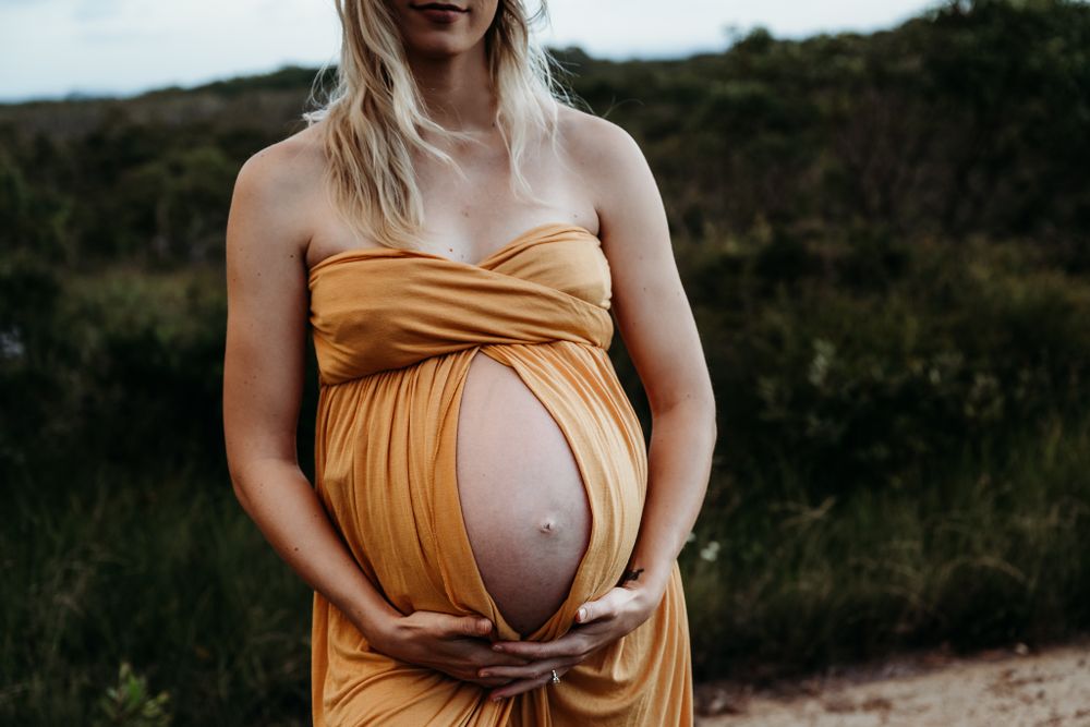 Person in mustard yellow dress displays baby bump against natural outdoor backdrop.