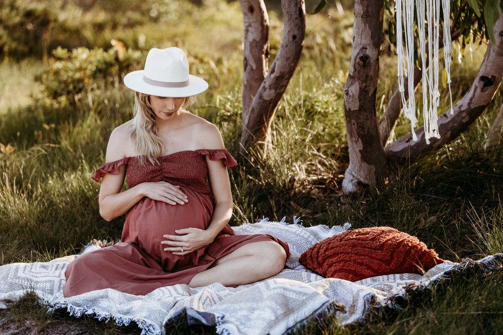 A woman in a flowy burgundy dress rests on a white blanket under a tree during a maternity photo session.