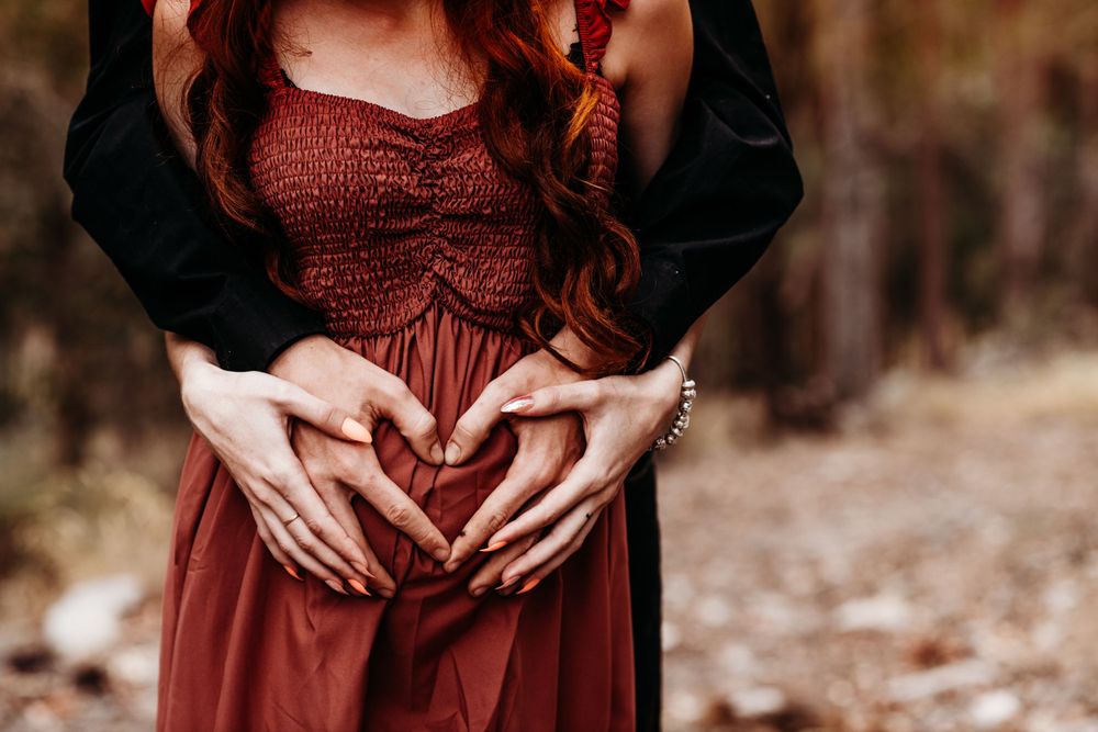 Hands forming heart shape over burgundy dress in romantic outdoor maternity photo.