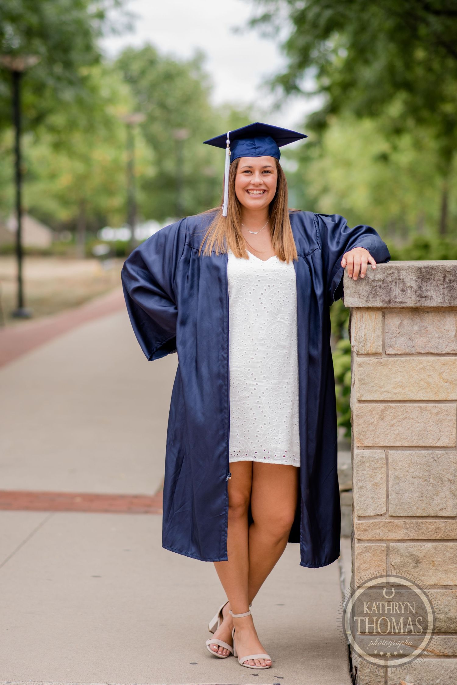 A graduate wearing blue cap and gown poses confidently against a stone wall on campus.