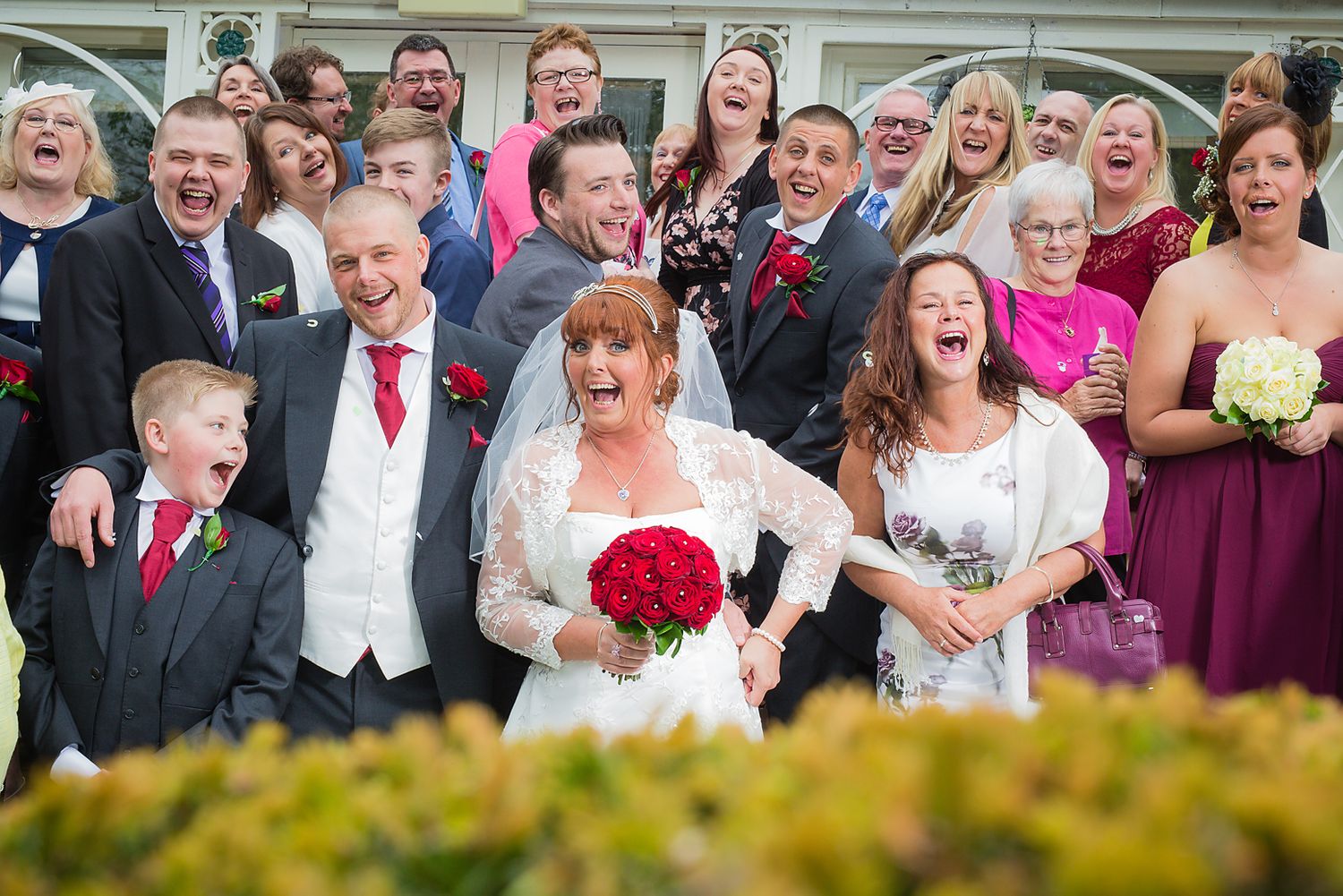 Happy group of smiling wedding guests gather with bride and groom outside Etrop Grange Hotel in Manchester
