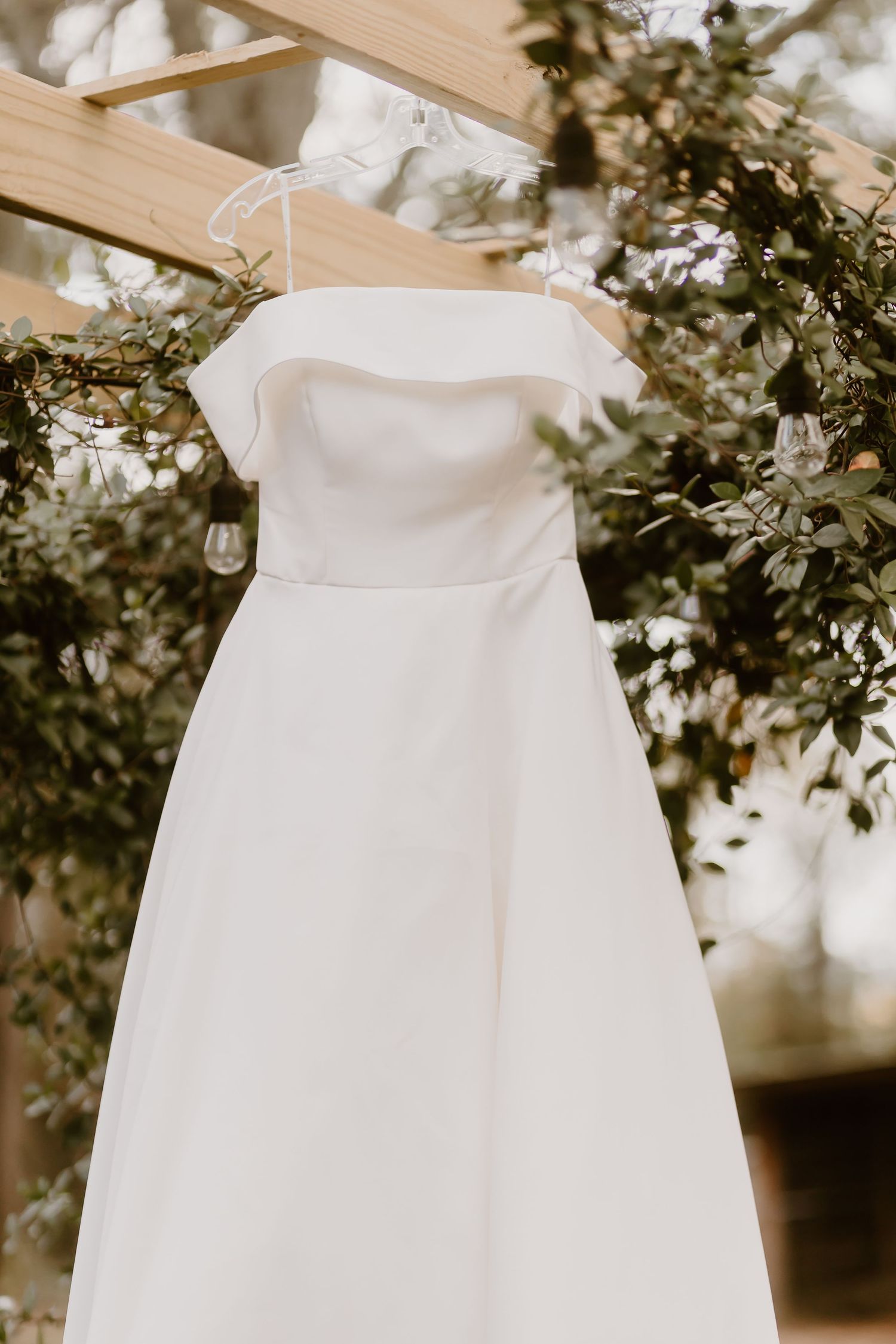 An elegant white wedding dress hanging on a wooden beam surrounded by greenery.