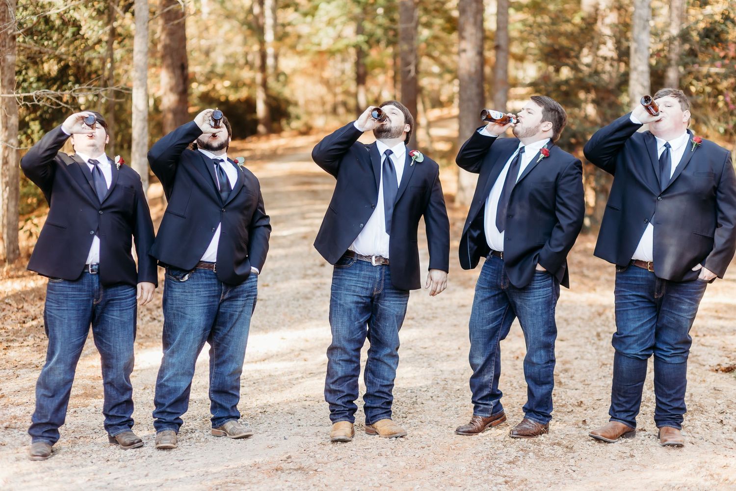 Groomsmen in navy suits and jeans pose playfully on a wooded path.