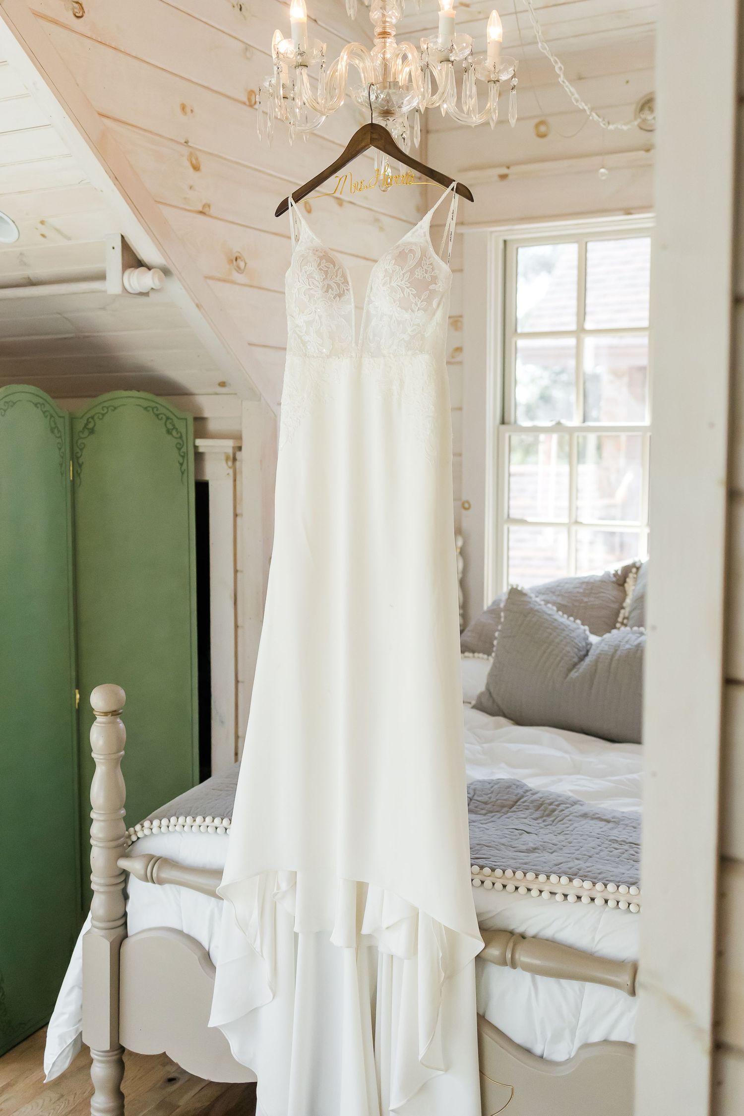 White wedding dress hanging from chandelier in bright, airy room with green accent furniture.