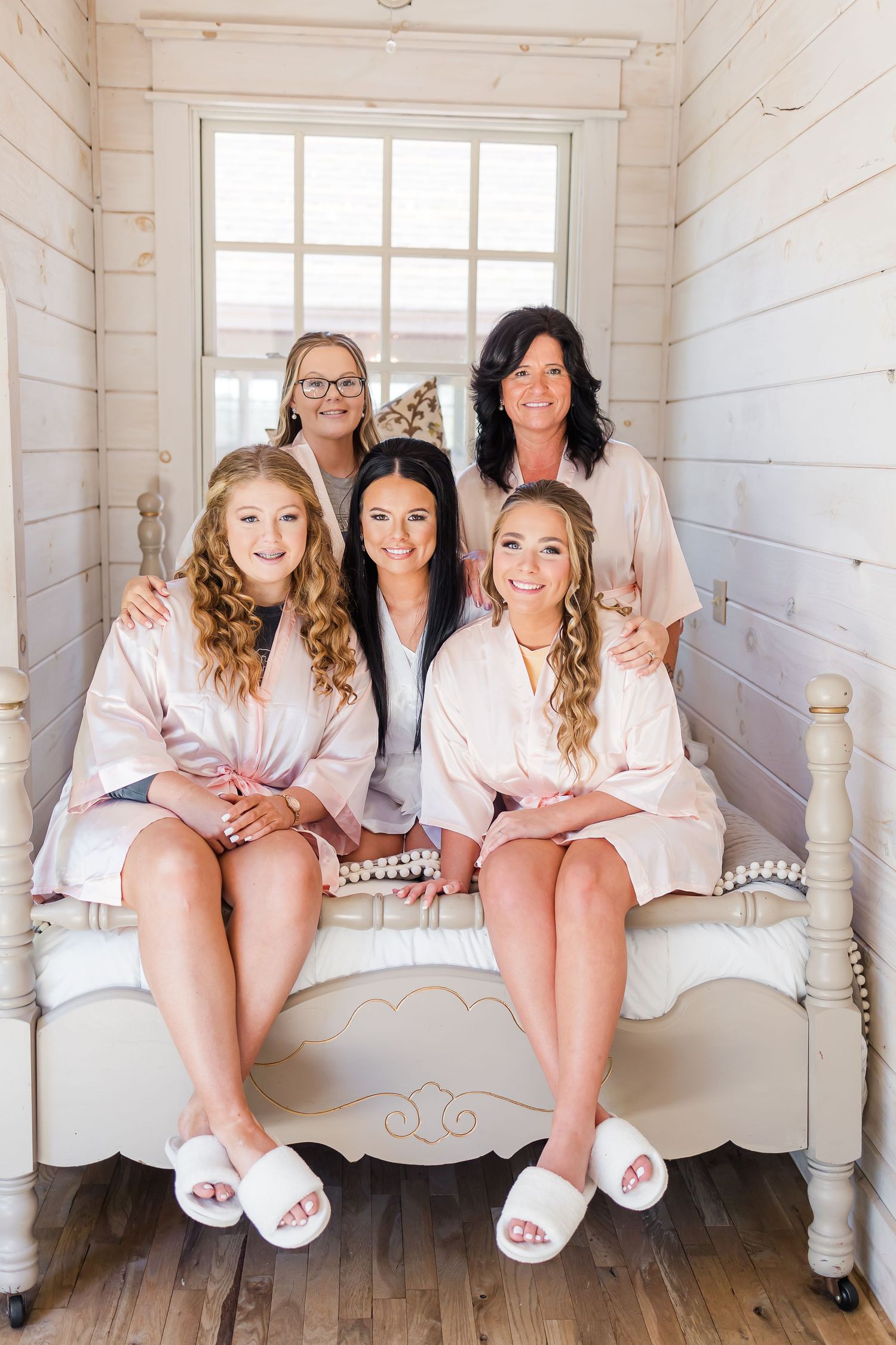 Group of bridesmaids in matching robes sitting on a white bench in a rustic room.