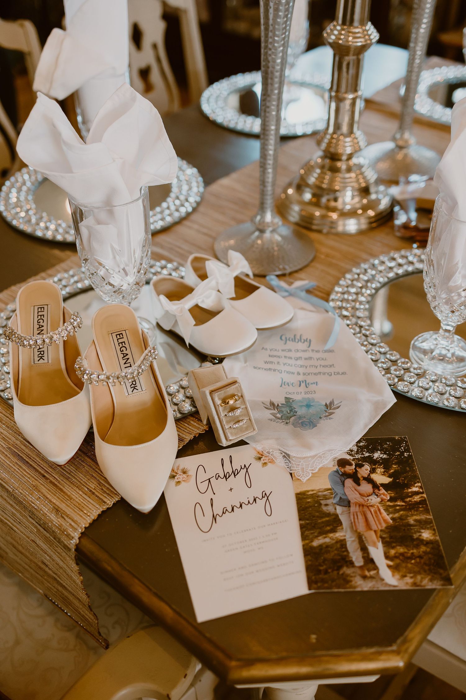 Wedding details including white shoes, invitation cards and table settings arranged on wooden surface.