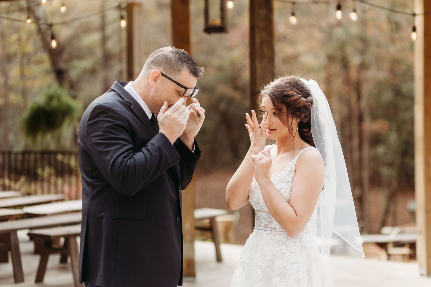 An emotional first look moment between newlyweds under string lights in a forest setting.