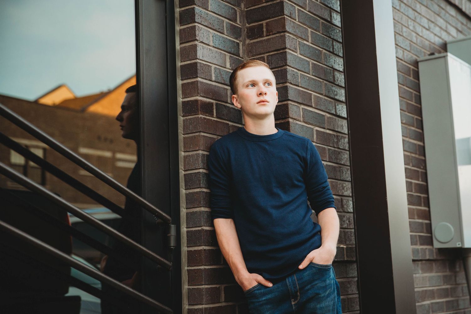 Senior boy in navy sweater poses against brick wall in urban setting.