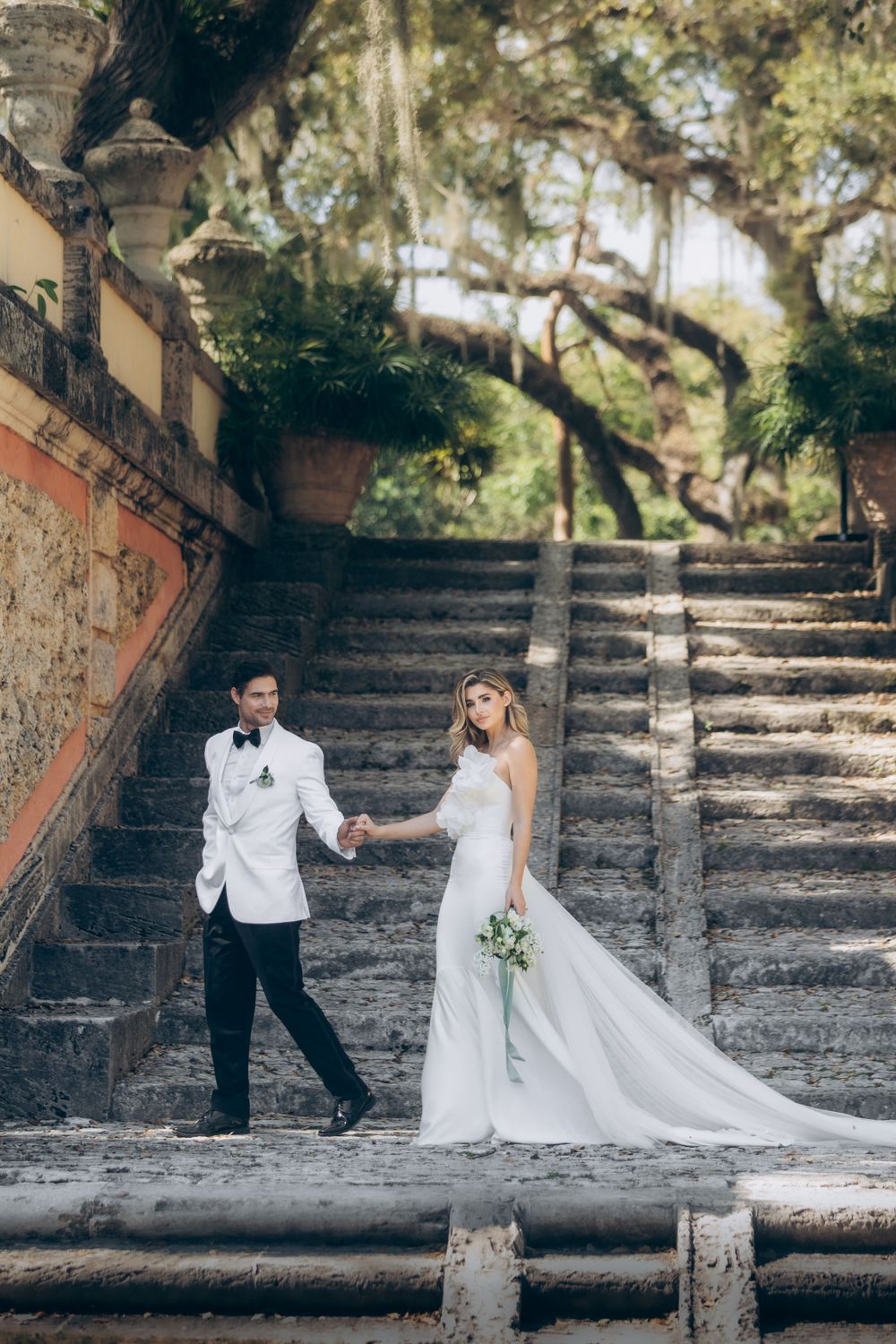 Newlywed couple descending stone steps under moss-draped trees.