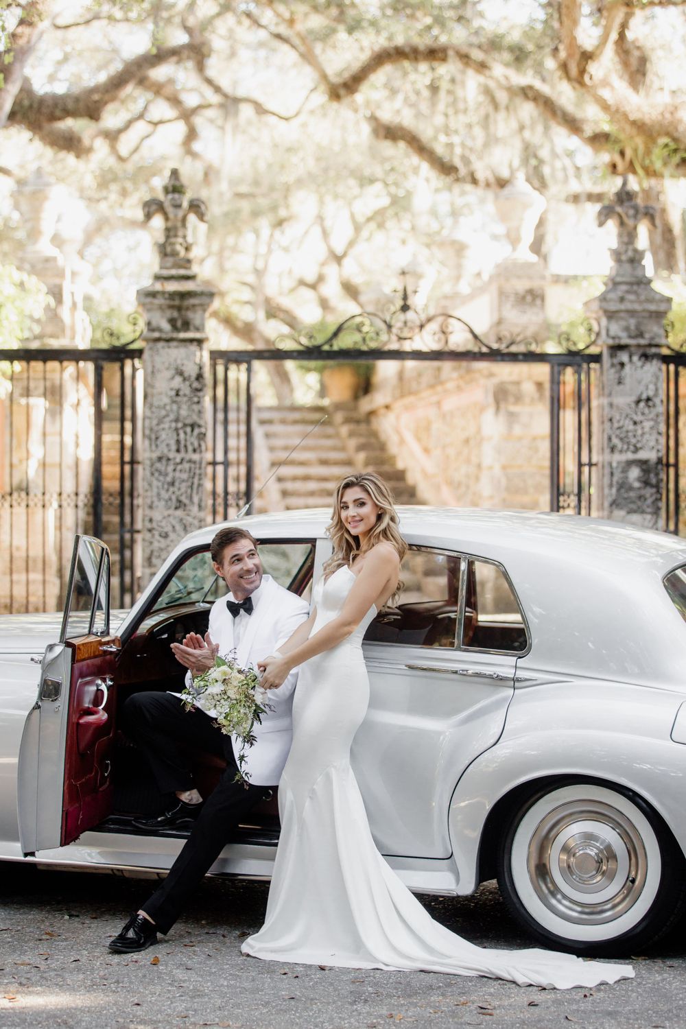 Elegant couple posing with vintage silver Rolls Royce at ornate gate entrance.