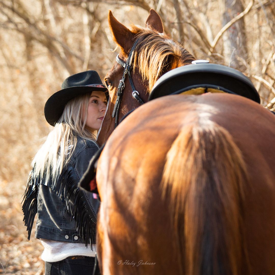A beautiful brown horse wearing a western saddle stands close to its rider in autumn light.