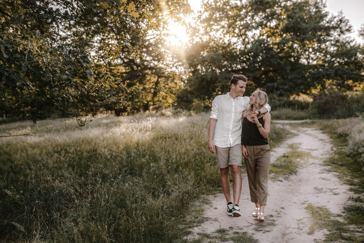 A couple walks together on a dirt path at sunset with trees and golden light in the background.