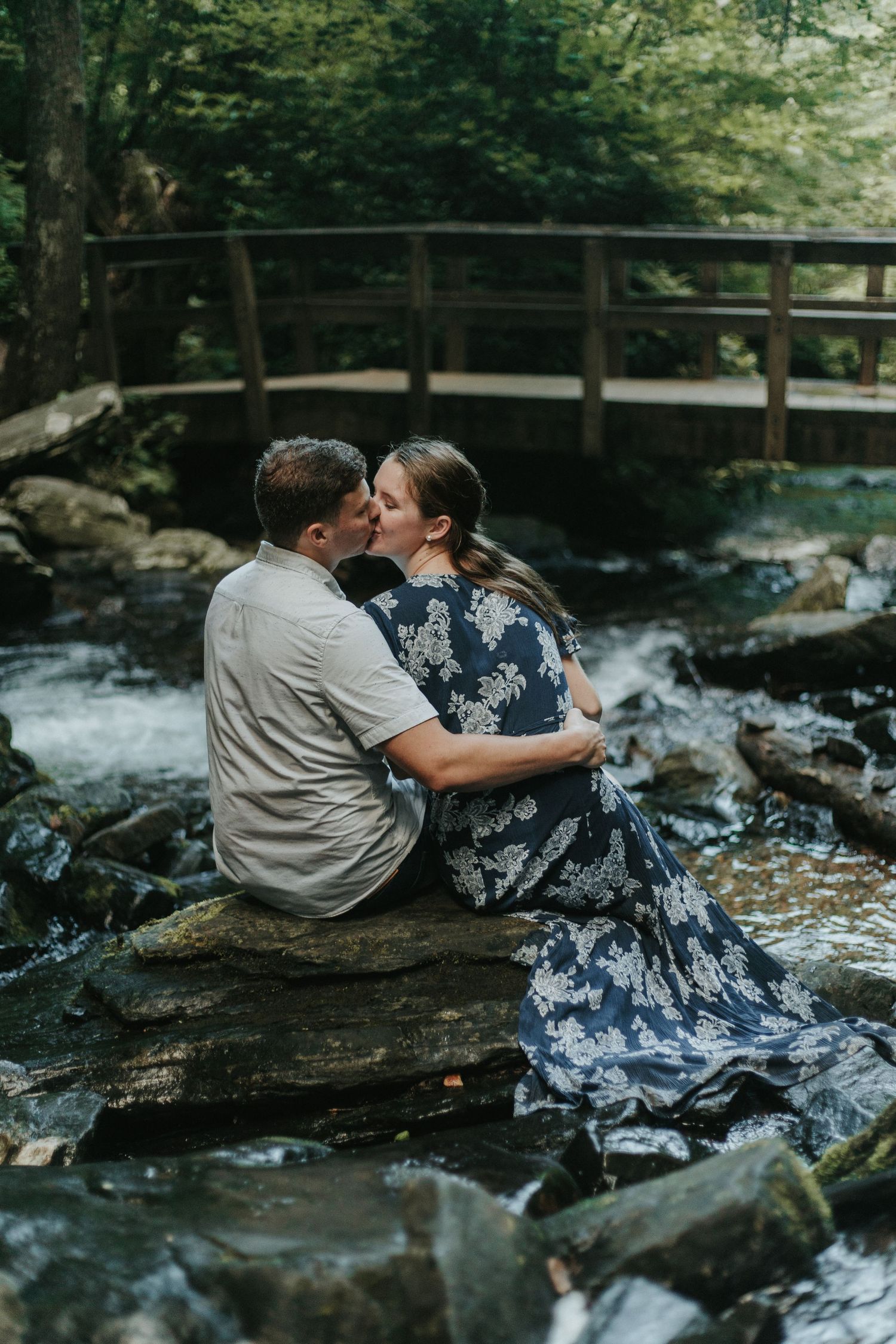 Romantic couple sitting on rocks by a stream with wooden bridge in background surrounded by forest scenery.