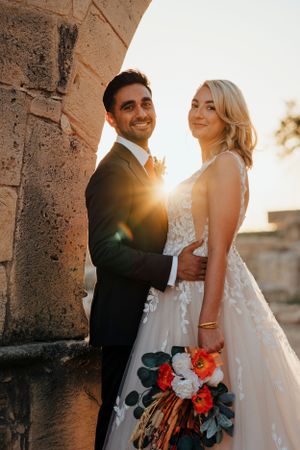 Newlyweds pose under stone arch during golden hour with vibrant coral bouquet.