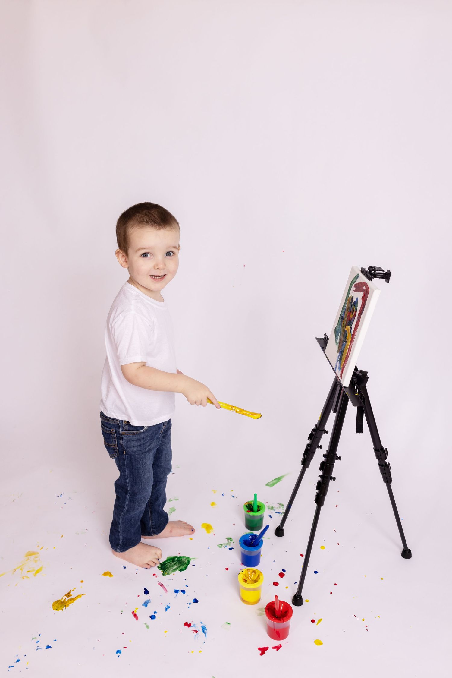 Young child painting at an easel with colorful paints scattered on white floor.